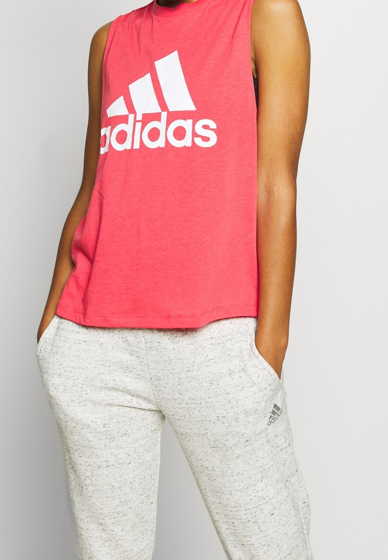 Personne portant un haut rouge sans manches Adidas et un pantalon de survêtement gris clair Adidas, les mains dans les poches sur un fond uni.