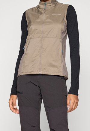 Patagonia vest in lichtbruin, met een hoge kraag, ritssluiting en blauwe ritsaccenten. Draagt over een zwart longsleeve shirt.