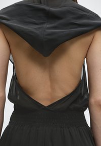 Οπίσθια όψη ατόμου που φοράει μαύρο αμάνικο φόρεμα με ανοιχτή πλάτη και μεγάλο τριγωνικό γιακά που καλύπτει τους άνω ώμους.