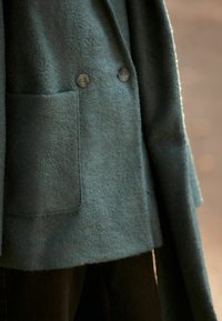 Zelený vlněný kabát s dvojitým zapínáním, dvěma velkými knoflíky a boční kapsou, s měkkým a texturovaným povrchem.