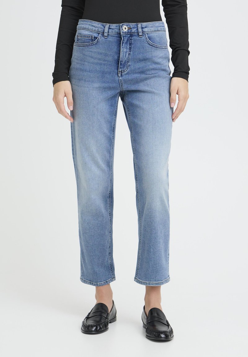 Osoba oblečená v svetlomodrých rifle s rovnými nohavicami a klasickým dizajnom päť vreciek, nosiaca čierne mokasíny, stojaca na bielom pozadí.