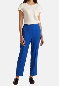 Pantalon bleu sur mesure en tissu lisse, avec une jambe droite, taille haute et poches latérales ; associé à un haut crème uni.