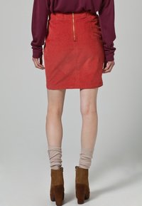 Minifalda de corduroy rojo con cierre de cremallera en la parte posterior, combinada con calentadores de piernas beige acanalados y botas de tacón ancho marrones.
