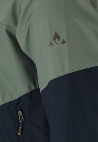 Vihreä ja tummansininen ulkoilutakin hiha, jossa on pieni geometrinen logo ja näkyvissä musta vetoketjullinen tasku.