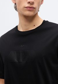 Νέος άνθρωπος που φοράει μαύρο T-shirt με διακριτικό ανάγλυφο λογότυπο "BOSS" και οριζόντια ρίγα στο στήθος.