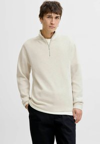 Svjetlosmeđi zip-up dukserica od teksturiranog materijala, dugih rukava i visokog ovratnika, uparenih s tamnim hlačama. Glatka površina.