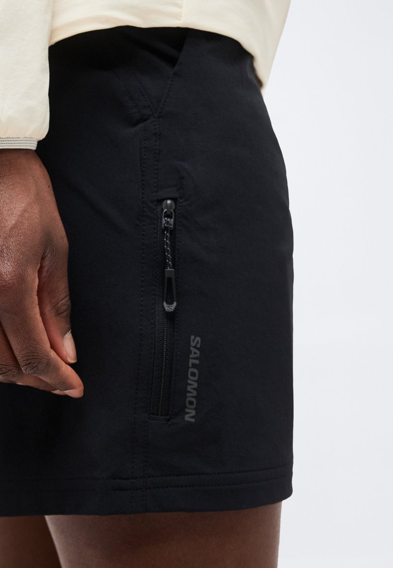 Gros plan sur la main d'une personne près d'une poche zippée noire Salomon sur un short noir avec une manche de couleur claire au-dessus.