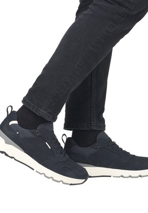 Marineblaue Sportschuhe mit einer strukturierten Oberseite, kontrastierender weißer Sohle und schwarzen Schnürsenkeln. Getragen mit schwarzer Jeans und Socken.