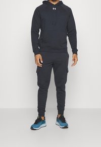 Hoodie bleu marine avec poche et capuche, associé à un pantalon de jogging assorti avec poches cargo. Porté avec des chaussures de course bleues et noires.