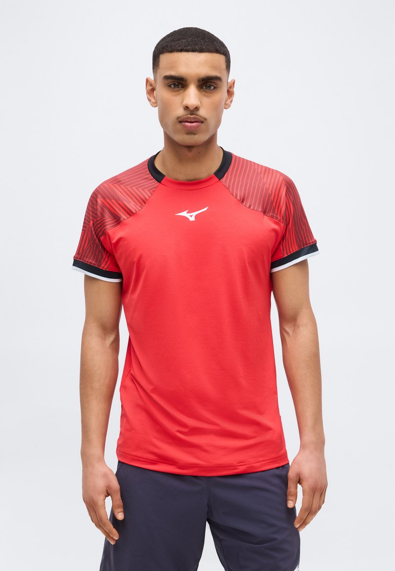 Νεαρός άνδρας με κοντά μαλλιά, φορώντας κόκκινο αθλητικό μπλουζάκι με μαύρα και κόκκινα μανίκια με σχέδιο και σκούρα σορτς, στέκεται μπροστά σε ένα αφαιρετικό φόντο.