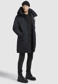 Joven con un gorro negro, un abrigo largo de invierno con capucha oscura, sudadera negra, jeans ajustados negros y botas gruesas negras, de pie frente a un fondo blanco.