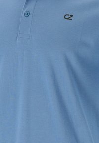 Vaaleansininen polo-paita, jossa on teksturoidusta kankaasta valmistettu pinta, kolmesta napista muodostuva kaulus ja pieni musta logo rinnassa. Pehmeä ja rento muotoilu.