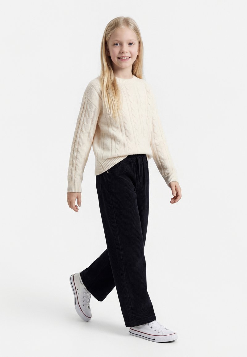 Noor tüdruk, kellel on pikkad blondid juuksed, naeratab, kõnnib kreemika palmikutega kampsunis, mustades laiade pükstega ja valgetes spordijalatsites valgel taustal.