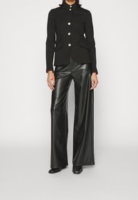 Chaqueta negra ajustada con botones plateados, cuello alto y bolsillos laterales, combinada con pantalones de cuero negros de pierna ancha y zapatos negros.