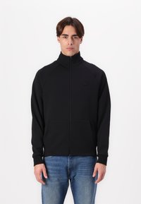 Veste zippée noire avec col haut, fabriquée en tissu doux, avec des manches raglan et deux poches avant. Logo Lacoste noir sur la poitrine.