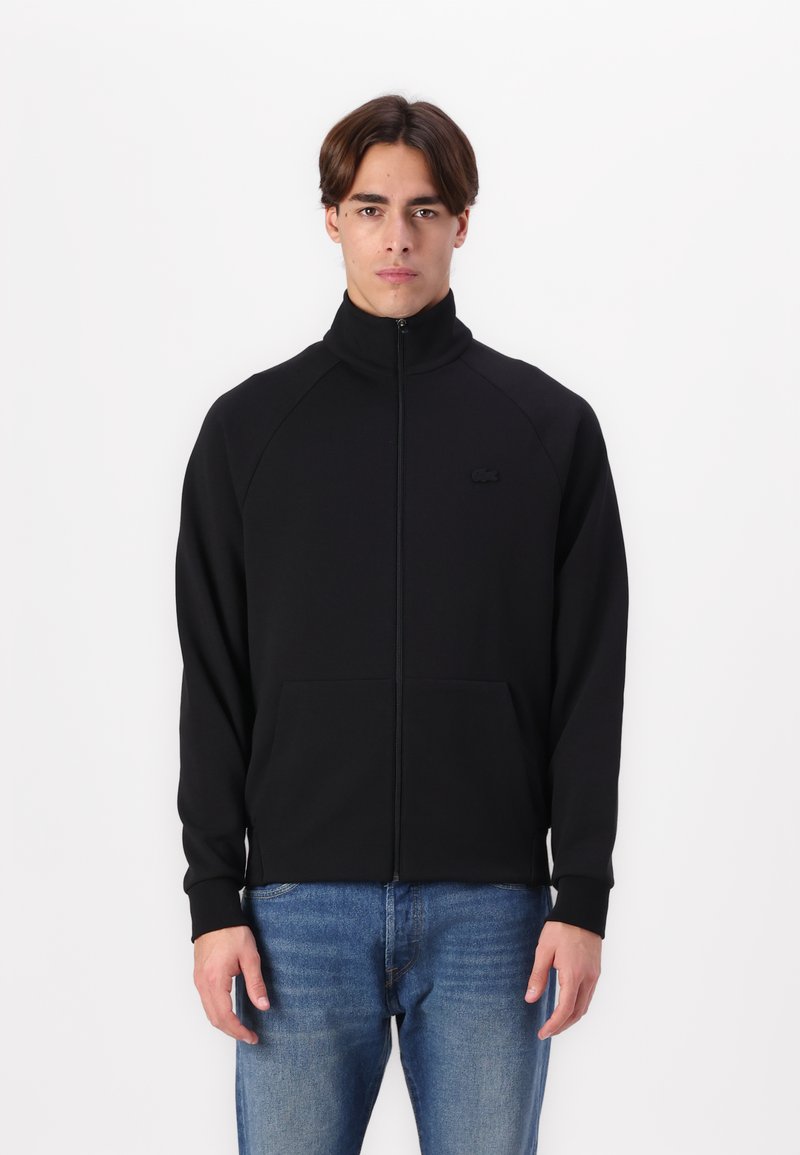 Veste zippée noire avec col haut, fabriquée en tissu doux, avec des manches raglan et deux poches avant. Logo Lacoste noir sur la poitrine.