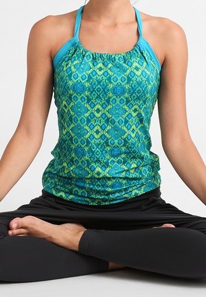 Persona che indossa un top senza maniche con motivo blu e verde e leggings neri, seduta a gambe incrociate in una posizione yoga su uno sfondo semplice.