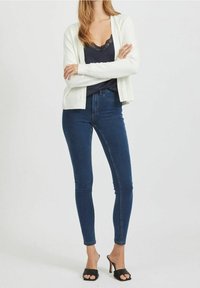 Kvinna klädd i en vit cardigan, en svart topp med spetsdetaljer, mörkblå tajta jeans och svarta klacksandaler, ståendes med armarna i kors.