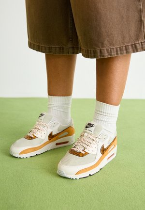 Jambes portant des baskets blanches Nike Air Max avec des touches orange, des chaussettes blanches et un short marron jusqu'aux genoux, debout sur un sol vert.