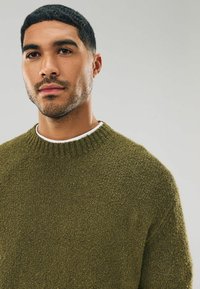 Olive green knit sweater with a textured finish, ribbed crew neckline, short sleeves, and a layered white shirt underneath.