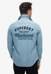 Jasnoniebieska koszula dżinsowa z pointed kołnierzykiem i zaokrąglonym dołem. Tył zdobi czarny haftowany napis: "SUPERDRY Precision Machined Copper Label."