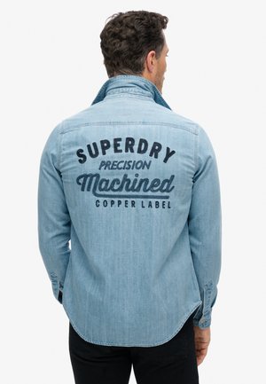 Jasnoniebieska koszula dżinsowa z pointed kołnierzykiem i zaokrąglonym dołem. Tył zdobi czarny haftowany napis: "SUPERDRY Precision Machined Copper Label."