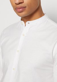 Gros plan sur une personne portant une chemise blanche à col mao avec des boutons sur le devant, ne montrant que les lèvres et le menton.