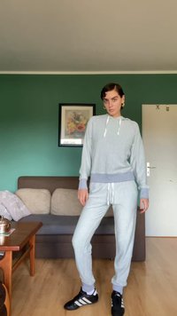 Světle šedá mikina s kapucí a odpovídající tepláky s kontrastními manžetami, šňůrkami a texturou. Černé sportovní boty s bílými pruhy.