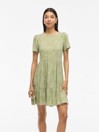 Abito verde a maniche corte con orlo a campana, caratterizzato da un motivo a pois e un corsetto aderente. Realizzato in tessuto leggero con una consistenza morbida.