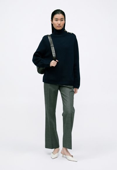 Marineblauwe oversized trui met hoge kraag en gouden knoopaccenten, gecombineerd met grijs geruite wijde broek en witte pointy-toe muilen.