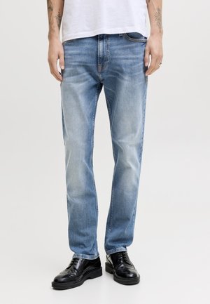 Voksen med lyseblå falmede straight-leg jeans, hvit skjorte, svarte skinn broguesko og synlige armtatoveringer.