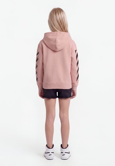 Child with long blonde hair wearing a pink hoodie with black sleeve designs, black denim shorts, and white Nike sneakers, facing away.
