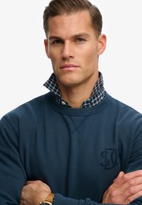 Felpa blu navy con collo a costine, indossata sopra una camicia a quadri con bottoni. Presenta un logo ricamato e dettagli di cucitura visibili.