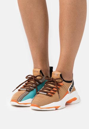 Pieds portant des baskets en tricot beige avec des accents orange et turquoise, des lacets noirs et orange, et des semelles blanches sur un fond blanc.