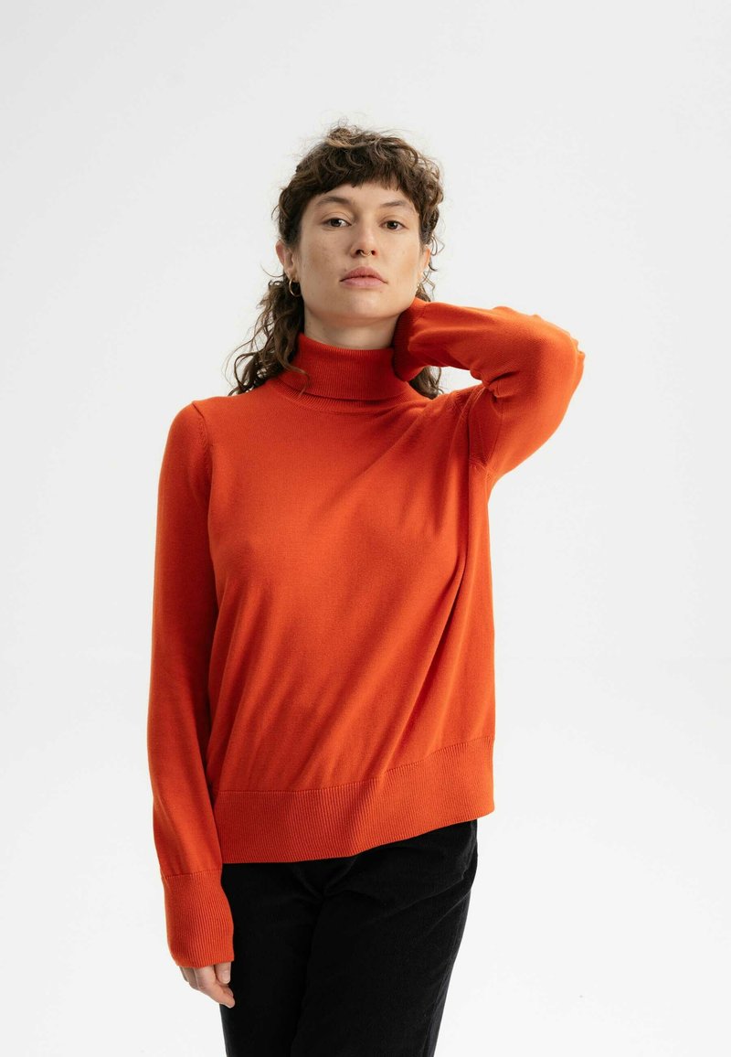 Roter Rollkragenpullover aus weichem Strick, mit langen Ärmeln sowie gerippten Bündchen und Saum. Getragen mit schwarzen Kordhosen.