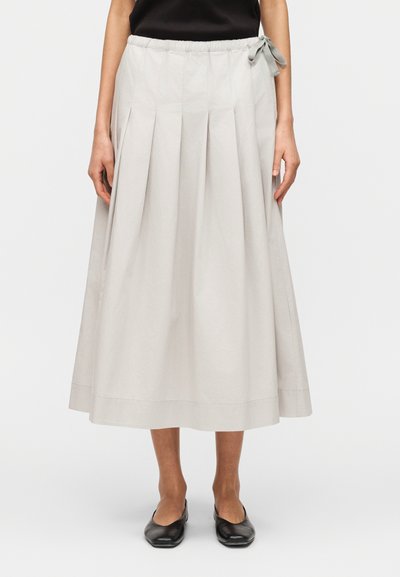 Femme portant une jupe midi gris clair à plis avec un nœud latéral, associée à des chaussures plates noires, debout devant un fond uni.