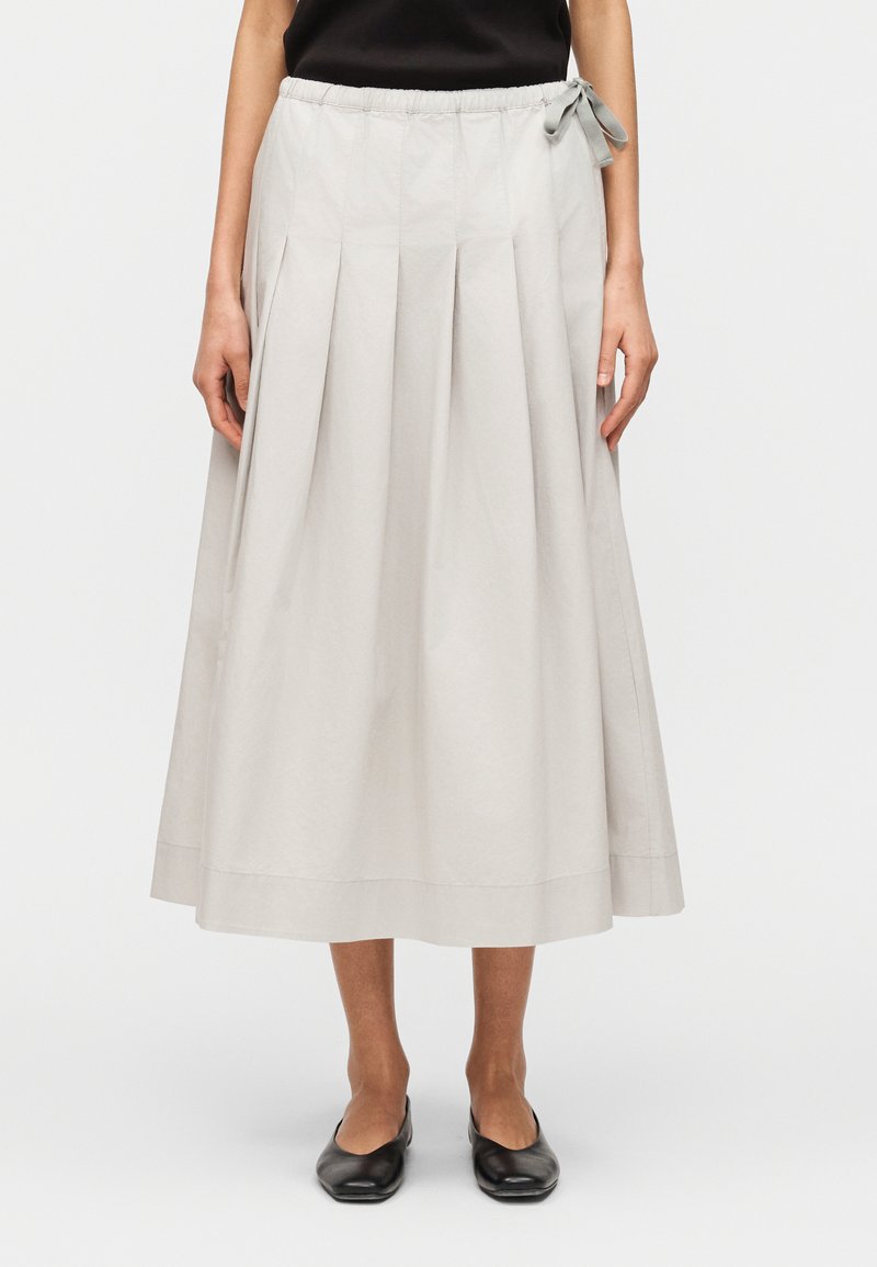 Γυναίκα με φωτεινή γκρι midi φούστα με πιέτες και πλευρικό φιόγκο, συνδυασμένη με μαύρα επίπεδα παπούτσια, στέκεται μπροστά σε απλό φόντο.