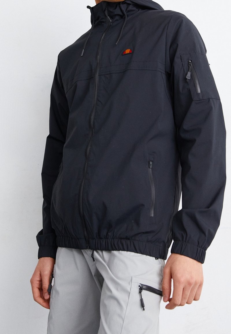 Homme portant une veste à capuche noire avec des poches zippées et un pantalon gris clair avec des poches latérales zippées.