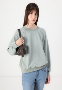 Felpa verde chiaro con scollo rotondo e maniche a raglan, abbinata a jeans blu. Borsa nera con tracolla e elementi decorativi.