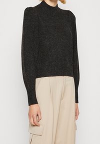 Osoba w ciemnoszarym swetrze z długim rękawem i beżowych spodniach o szerokich nogawkach z bocznymi kieszeniami cargo.