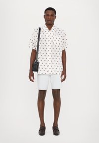 Cămașă albă cu mânecă scurtă și model floral; asociată cu pantaloni scurți din denim deschis și loafers negri; purtând o geantă mică neagră.