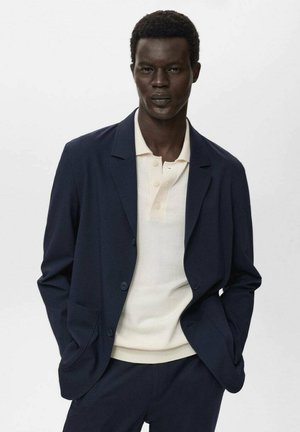 Navy blue tailored blazer with notch lapels, paired with a cream short-sleeve polo shirt. Slim fit with minimal detailing, smooth fabric texture.
