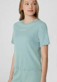 Camiseta de rayas verde claro con mangas cortas, que presenta puños y dobladillo con volantes, y texto bordado "Lovely" en el frente.