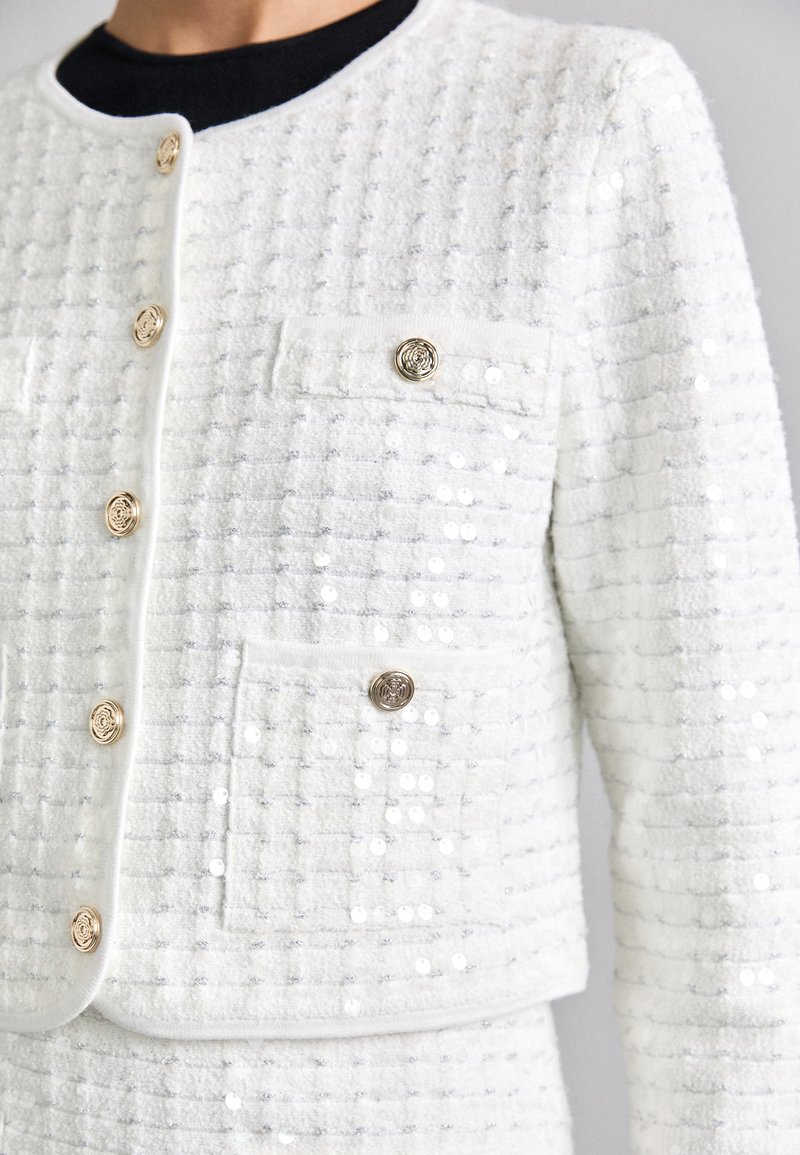 Veste blanche texturée avec des accents de boutons en or, deux poches avant et une coupe ajustée. Le tissu a un léger éclat et un design structuré.