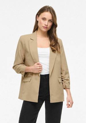 Hosszú barna hajú nő bézs zakót visel fehér felső és fekete nadrág fölött, egyszerű fehér háttér előtt áll.