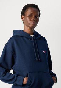 Persona con capelli corti e ricci che indossa una felpa con cappuccio blu navy con un piccolo logo Tommy Jeans, mani nella tasca frontale, sfondo neutro.