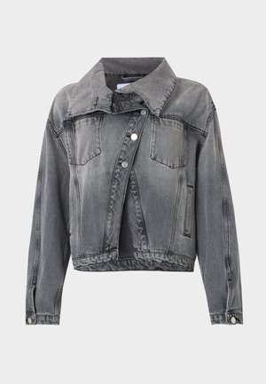 Zwart persoon die een oversized vervaagde grijze spijkerjas draagt, wit t-shirt, donkere uitlopende spijkerbroeken, zwarte hakken en zwarte zonnebril, staand.