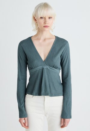 Teal langærmet bluse med tekstureret stof, V-halsudskæring og peplumdesign. Har en tætsiddende talje og en svajende bund.