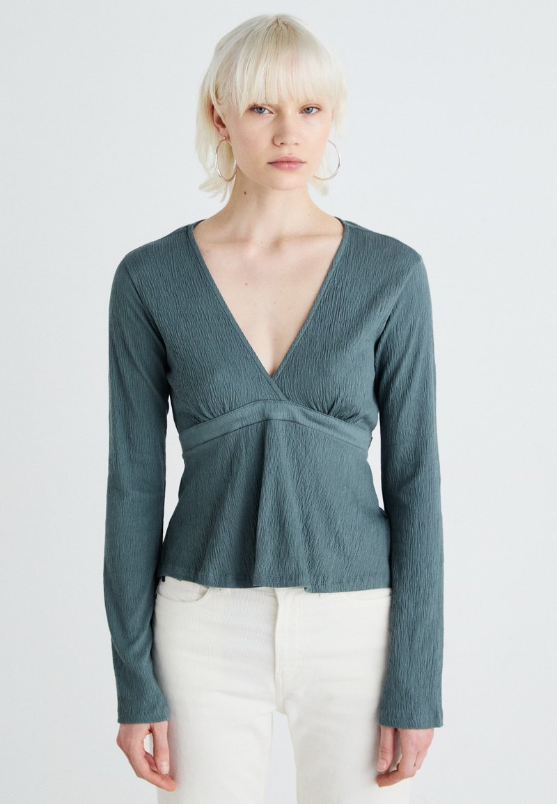 Turkos långärmad topp med texturerad tyg, V-ringning och peplumdesign. Har en figurnära midja och ett flödande underparti.