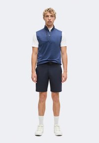 Jeune homme portant un gilet sans manches bleu zippé sur un t-shirt blanc à manches courtes, un short bleu marine, des chaussettes blanches et des baskets blanches, debout devant un fond uni.